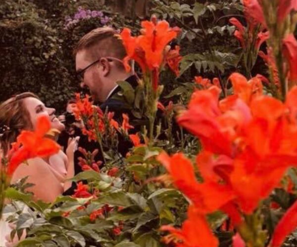 A wedding couple staring at each other behind a bunch of orange flowers